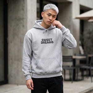 Puede incluir: Una sudadera con capucha gris claro con el texto "SWEET DREAMS" en negro. La sudadera tiene una capucha con cordón y un bolsillo delantero. La persona de la imagen lleva pantalones negros.