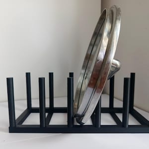 May include: A black metal rack with multiple vertical pegs designed for organising kitchen items. A single circular silver metal pot lid with a central handle is positioned upright between the pegs of the rectangular storage organiser.