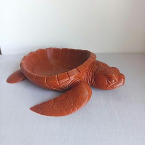 Op de afbeelding: Een terracotta-kleurige kom in de vorm van een schildpad met een gedetailleerd schildpatroon dat de kom vormt. De schildpad heeft een gebeeldhouwde kop, vier flippers en een textuur die het uiterlijk van schubben over het hele lichaam nabootst.