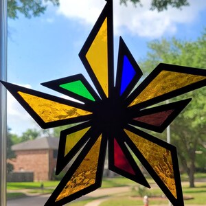 May include: A colorful stained glass starburst suncatcher. The starburst features a black outline with yellow, green, blue, and red glass panels. The suncatcher is hanging in front of a window with a view of a neighborhood.