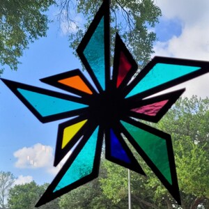 May include: A colorful stained glass star suncatcher with a black outline. The star features various colored glass pieces, including turquoise, orange, red, yellow, blue, and green. The suncatcher hangs in front of a blue sky with trees.