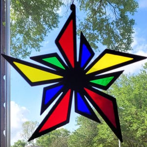 May include: A colorful stained glass starburst suncatcher. The starburst features red, yellow, blue, and green geometric shapes outlined in black. The suncatcher is hanging in front of a window with a view of trees and a blue sky.