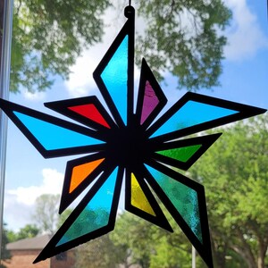 May include: A colorful stained glass star with a black outline. The star features various geometric shapes in shades of blue, red, orange, yellow, green, and purple. The star is hanging in front of a window with a view of trees and a blue sky.
