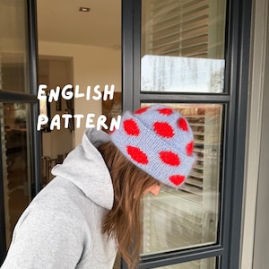 Könnte beinhalten: Hellblaue Strickmütze mit roten Punkten. Der Text "ENGLISH PATTERN" ist in Weiß zu sehen. Die Person trägt einen grauen Kapuzenpullover.