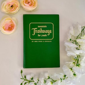 May include: A green hardcover book titled Mountain Trailways for Youth by Mrs. Chas. E. Cowman, featuring gold lettering and a gold-bordered rectangular frame on the front cover, surrounded by pale pink ranunculus and white floral stems.