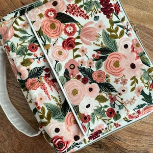 May include: A floral-patterned book cover and matching case. The fabric features a white background with pink, red, and burgundy flowers, green leaves, and a gold zipper. The case has a white handle and trim.