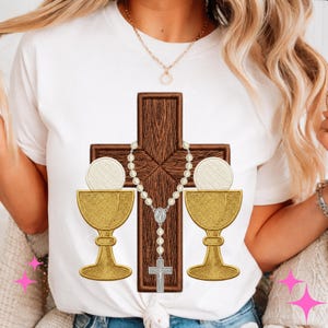Può includere: T-shirt bianco con un ricamo a tema religioso. Il disegno include una croce di legno marrone, un rosario, due calici dorati e due ostie bianche. Il disegno è centrato sulla parte anteriore della maglietta.