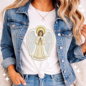 May include: White t-shirt featuring an embroidered angel design with a gold halo and wings. The angel is depicted with folded hands and a flowing gown. The shirt is paired with a denim jacket and jeans.