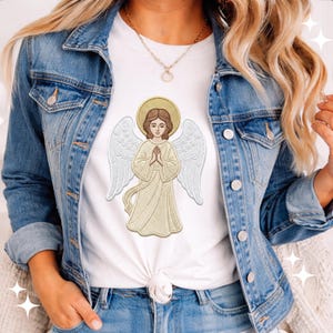Peut inclure: T-shirt blanc orné d'un motif brodé d'un ange avec une auréole et des ailes dorées. L'ange est représenté les mains jointes en prière. Le t-shirt est associé à une veste en jean et un jean.