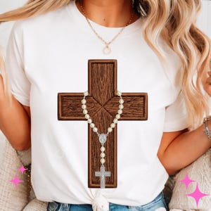 May include: White t-shirt featuring a detailed embroidered cross with a rosary. The cross is a dark brown color with a wood-grain texture. The rosary has white beads and a silver-colored crucifix and centerpiece.