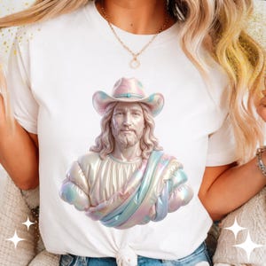 Puede incluir: Camiseta blanca con una representación 3D de una figura que lleva un sombrero de vaquero y un chal colorido. La figura tiene pelo largo y barba. El sombrero y el chal tienen colores pastel. La camiseta está atada a la cintura.