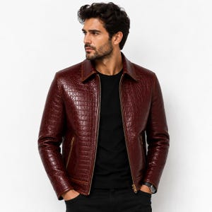 Könnte beinhalten: Eine dunkelburgunderfarbene Jacke mit texturiertem Krokodilledermuster, klassischem Kragen und durchgehendem Frontreißverschluss. Das Kleidungsstück wird über einem schlichten schwarzen Shirt getragen und hat lange Ärmel mit Bündchen und zwei Seitentaschen.