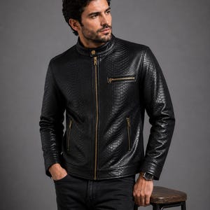 Puede incluir: Una chaqueta negra con un patrón texturizado de piel de serpiente, que presenta un cuello alzado con un botón a presión dorado, una cremallera frontal completa y tres bolsillos con cremallera y herrajes dorados en el pecho y la cintura.
