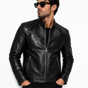 Könnte beinhalten: Eine schwarze Jacke mit strukturiertem Schlangenledermuster, ausgestattet mit einem Stehkragen, einem durchgehenden Frontreißverschluss und zwei seitlichen Reißverschlusstaschen. Das Kleidungsstück wird über einem schwarzen Shirt getragen.