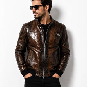 Puede incluir: Una chaqueta marrón con textura de piel de serpiente, cremallera frontal plateada y puños acanalados negros en la cintura y las muñecas. La chaqueta presenta un pequeño emblema metálico plateado en el lado izquierdo del pecho.