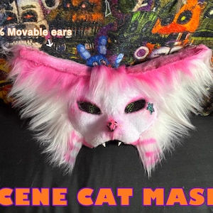Op de afbeelding: Een roze en wit kattenmasker met pluizig haar en gaasbedekkingen voor de ogen. Het masker heeft beweegbare oren en de tekst "SCENE CAT MASK!" wordt weergegeven in oranje letters.