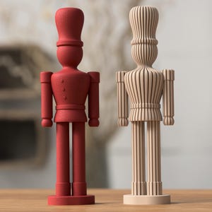 Könnte beinhalten: Zwei dekorative Holzsoldatenfiguren. Eine ist einfarbig rot mit strukturierter Oberfläche, die andere ist aus Naturholz mit Streifendesign. Beide haben hohe Hüte und stehen aufrecht.