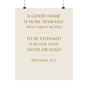 Peut inclure: Une affiche avec le texte "A GOOD NAME IS MORE DESIRABLE THAN GREAT RICHES. TO BE ESTEEMED IS BETTER THAN SILVER OR GOLD. PROVERBS 22:1" en or sur un fond crème. L'affiche est maintenue par deux clips noirs.