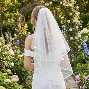 Puede incluir: Un velo de novia blanco con un diseño en capas y ribete de satén. El velo lo lleva una persona con un vestido blanco, de pie en un jardín con flores y vegetación.