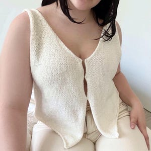 May include: Cream-colored, sleeveless knit top with a deep V-neckline. The top has a split front design and a textured knit pattern. The garment is likely handmade and suitable for warm weather. The top is a neutral color, making it versatile for various outfits.