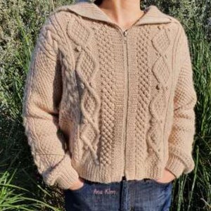 Puede incluir: Un suéter beige con cremallera y capucha, tejido con trenzas. El suéter presenta intrincados patrones de cable y dobladillos y puños acanalados. La cremallera recorre el centro y el suéter se lleva sobre unos vaqueros azules.