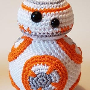 May include: A handmade crochet BB-8 droid. The spherical body is white with orange and gray accents. The head is white with orange and gray stripes, and it has two black button eyes. The droid is a popular character from the Star Wars franchise.