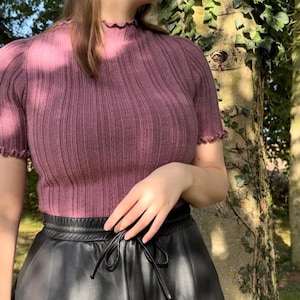 May include: A mauve-colored ribbed knit top with short, ruffled sleeves and a matching ruffled neckline. The top is paired with a black leather-look skirt with a drawstring waist. The background features a tree trunk with green ivy.