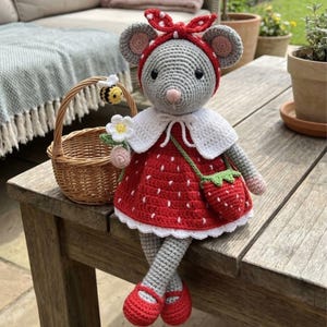 Op de afbeelding: Gehaakte muispop in een rode aardbeienjurk, rode schoenen en een polkadot haarband. De pop heeft een witte kraag, een groene aardbeientas en houdt bloemen vast. Een klein mandje met een bij staat ernaast.