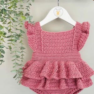 Puede incluir: Un pelele de ganchillo rosa empolvado con volantes y botones de madera en forma de corazón. La prenda para bebé se exhibe en una percha blanca, mostrando la artesanía hecha a mano. El pelele presenta un escote cuadrado y mangas cortas.