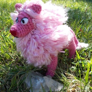 Puede incluir: Un león rosa hecho a mano con ganchillo, con una melena y cola esponjosas, de pie en hierba verde. El león tiene ojos grandes y una nariz roja, mostrando un diseño caprichoso. Este peluche es un artículo único y artesanal.
