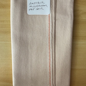 May include: A folded piece of beige Aida fabric with a visible grid weave. A white tag is pinned to the top, featuring the handwritten text: 16 CT ANTIQUE MUSHROOM FAT QTR. The fabric edge shows a distinct orange vertical line along the selvage.