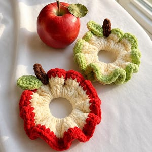 May include: Two crocheted hair scrunchies shaped like apple slices, one with red edges and one with green edges, both featuring a cream-colored center, a small green leaf, and a brown stem, positioned next to a fresh red apple.