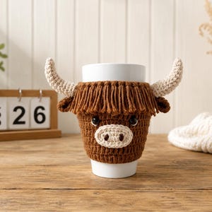 May include: A white ceramic mug featuring a brown crocheted cozy shaped like a Highland cow. The cozy includes a fringed brown mane, two light beige horns, and a light beige snout with dark eyes, fitting snugly around the cylindrical mug.