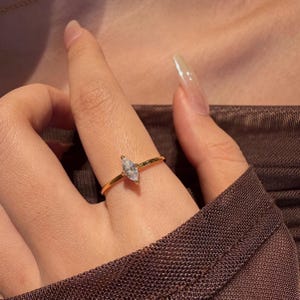 May include: A delicate gold ring featuring a marquise-cut diamond. The ring's slender band complements the sparkling gemstone, creating a refined and elegant piece of jewelry. The ring is shown on a finger.