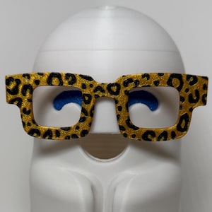 Pode incluir: Uma estatueta de fantasma branca com acabamento fosco liso, apresentando uma cabeça arredondada e corpo drapeado. O fantasma usa óculos de aro grosso com estampa de leopardo dourada e preta e dois pequenos detalhes circulares azuis.