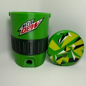 May include: A bright green cylindrical cup featuring the Mtn Dew logo in red, white, and black. The cup has a black textured band around the middle and a small circular opening at the base. A matching circular lid with a geometric pattern is included.