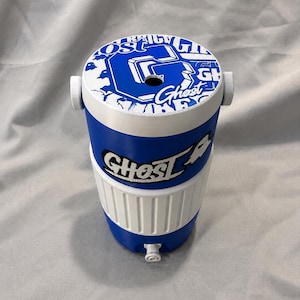 May include: A blue and white cooler with the word "Ghost" and other graphics. The cooler has a spigot and two white handles. The top of the cooler has a blue and white design with the word "Ghost" and other graphics.