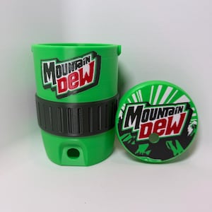 May include: A bright green plastic cup with a black textured grip band and a matching circular lid. Both the cup and the lid feature the Mountain Dew logo in white, red, and black. The cup has a small circular opening at the base and a straw hole.