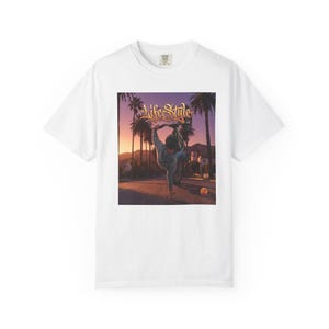 Puede incluir: Una camiseta blanca con un estampado gráfico de una persona haciendo el pino sobre una bicicleta en medio de una calle bordeada de palmeras al atardecer. El texto "Life Style" aparece impreso en una fuente cursiva dorada encima.