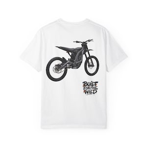 Puede incluir: Una camiseta blanca de manga corta con un gráfico negro de una moto de cross eléctrica en la parte posterior. Debajo de la moto, el texto BUILT FOR THE WILD está impreso en una fuente negra estilizada con detalles en naranja, como barro.