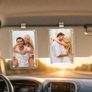 May include: Two clear acrylic photo frames with silver clips hang from a car visor. Each frame displays a color photograph of a couple. The photos are attached with silver screws. The background shows a blurred road.