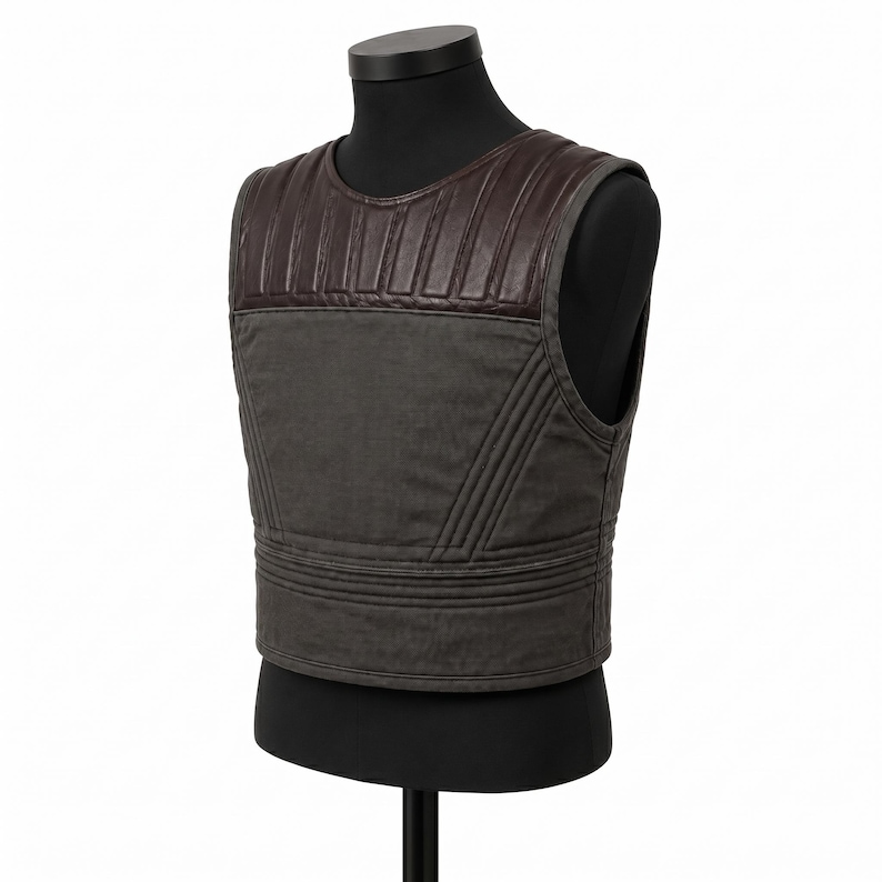 May include: A dark gray vest with a brown leather upper section. The vest has a quilted design on the shoulders and horizontal stitching across the lower section. It is displayed on a black mannequin.