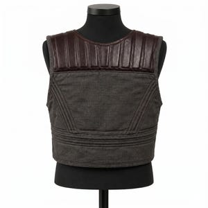 May include: A gray vest with a brown leather shoulder panel. The vest has a quilted design and is displayed on a black mannequin. The vest is a neutral color and could be worn by anyone.