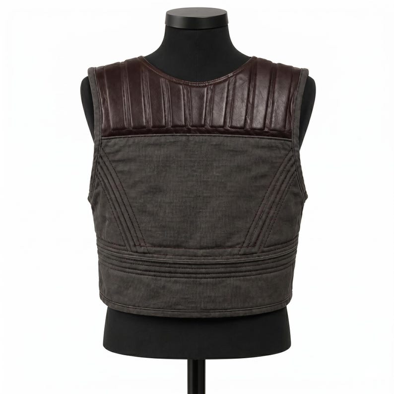 May include: A gray vest with a brown leather shoulder panel. The vest has a quilted design and is displayed on a black mannequin. The vest is a neutral color and could be worn by anyone.