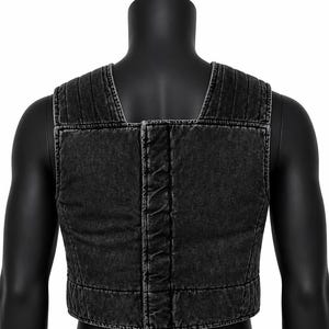 May include: A black denim vest with a square neckline and a cropped fit. The vest features vertical stitching on the shoulders and a central seam down the back. The fabric has a faded, worn appearance.