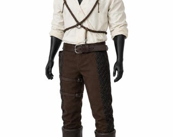 May include: A cream-colored long-sleeve shirt with a V-neck, worn with a brown leather harness featuring a central ring. The outfit includes dark brown trousers with quilted side panels, two side straps with buckles, a leather belt, and dark brown boots.