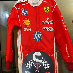 May include: Red racing suit with white detailing, displaying sponsor logos including HP, Shell, and IBM. The suit features a Ferrari emblem and the text "RICHARD MILLE". A black circular sticker with a helmet and checkered flags is attached.