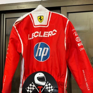 May include: Red racing suit with white detailing, displaying the Ferrari logo and the name "LECLERC". The suit includes the HP logo and the text "RICHARD MILLE". A circular sticker with a helmet and checkered flags is also visible.