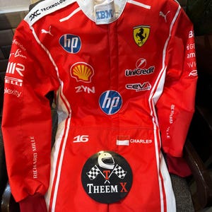 May include: A red and white racing suit with various sponsor logos, including "HP," "Shell," and "Unicredit." The Ferrari logo, the number "16," and the name "CHARLES" are displayed. The text "THEEM X" is also visible.