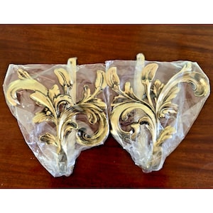 May include: Two gold-toned metal curtain tiebacks featuring an ornate, symmetrical leaf and scrollwork design. Each piece is individually encased in a clear plastic protective sleeve, showcasing the metallic finish and detailed relief patterns.